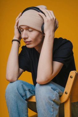 Sıradan giyinmiş, kulaklık takmış, parlak bir arkaplanda düşünceli bir poz veren genç bir adam.