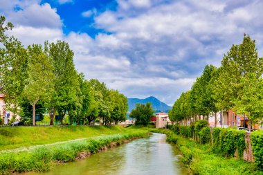 The Topino River flowing through Foligno, Italy, surrounded by green trees and a mountainous backdrop. clipart