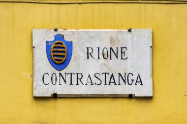 İtalya 'nın Umbria kentindeki Foligno kentindeki Rione Contrastanga' nın mermer plaketinde mavi arka planda üç altın külçeden oluşan bir amblem yer alıyor.. 