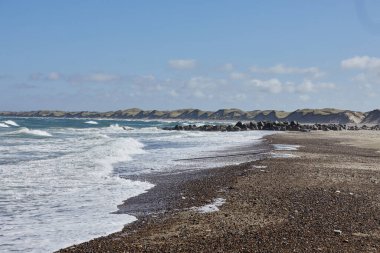 Kumsal ve deniz dalgaları sahile vuruyor. Arka planda kara, kum tepeleri ve som kayaları var. Güneş parlıyor ve gökyüzü mavi..