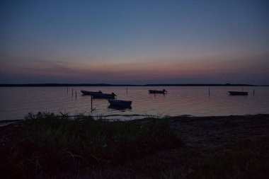 Alacakaranlıkta botlar suyun üzerinde. Ön planda kumsal var. Gökyüzü ve su mavi ve Rosa renginde. Dikey.