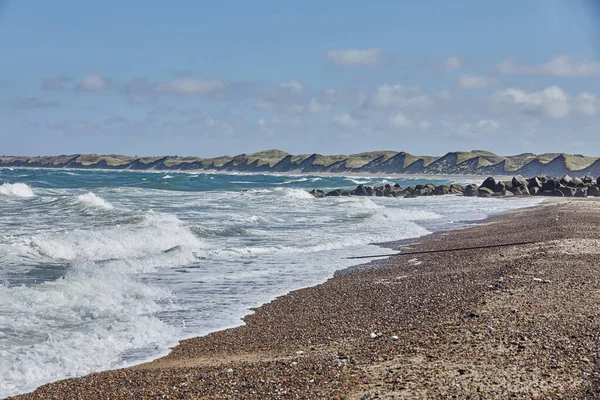 Kumsal ve deniz dalgaları sahile vuruyor. Arka planda kara, kum tepeleri ve som kayaları var. Güneş parlıyor ve gökyüzü mavi.