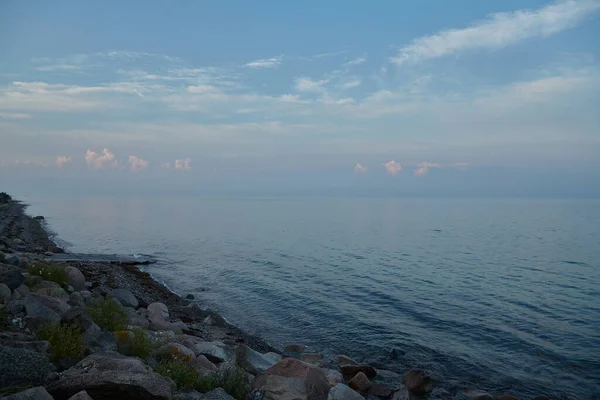 Gün batımında Sean ve plajın manzarası. Gökyüzü küçük beyaz bulutlarla mavidir. Ön planda büyük kayalar var..