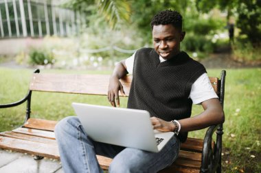 Yelekli genç adam dışarıda dizüstü bilgisayarda çalışıyor, ekrana bakıyor, gülümsüyor. Üniversite kampüsünde bankta oturan öğrenci, uzaktan kumandalı işten ya da uzaktan eğitimden keyif alıyor.
