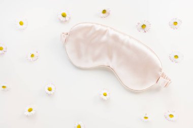 Pembe saten göz uyku maskesi ve taze bahar veya beyaz arka planda papatya çiçekleri. Uykusuzluk, uykusuzluk, güzellik uykusu konsepti. Kızlar ve kadınlar için aksesuarlar