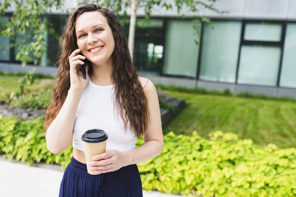 年轻女子笑着喝咖啡 坐着纸杯走出去 用手机聊天 走在城市街道上 春夏季节 白色作物顶面的模型 图库照片