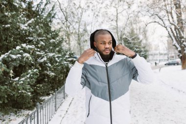 Young African man in casual white and gray clothes, sport jacket with hood . Winter season street portrait in park with snow. Lifestyle, city life concept