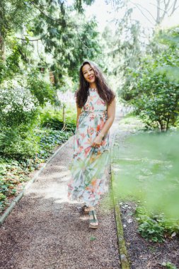 Beautiful happy multiracial Japanese woman in romantic floral maxi dress in summer park, smiling. Full body shot. Sunlight, copy space