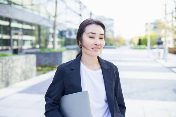 Relaxed and confident Asian business woman in suit holding laptop, outdoor. Job, work aspirational concept, background of office center. Business people lifestyle, copy space