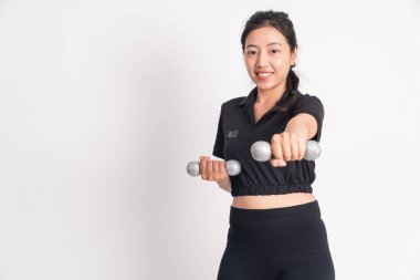 Halteri tutan gülümseyen kadın ısınırken arka planda hafif ağırlıklar kaldırıyor.