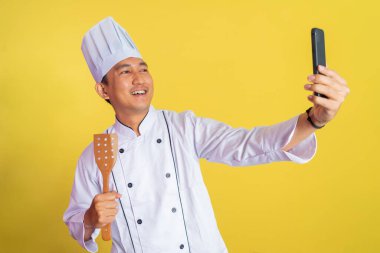 Asyalı erkek şef, arka planda spatula tutarken şef ceketi giyiyor.