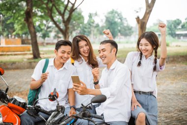 Dört mutlu lise öğrencisi motosikletle mezuniyeti kutlamak için akıllı telefonlar kullanıyor.