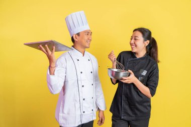 Erkek aşçı ekmek tepsisi taşıyor ve kadın aşçı çırpıcıyla karıştırma kabını kullanıyor.