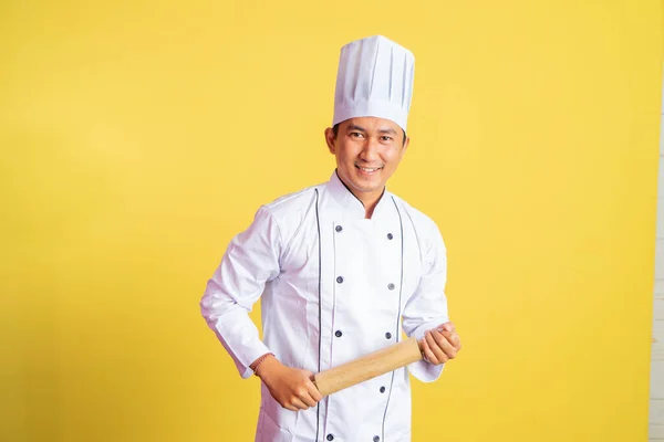 Asian chef smiling at camera while holding wooden rolling pin on isolated background