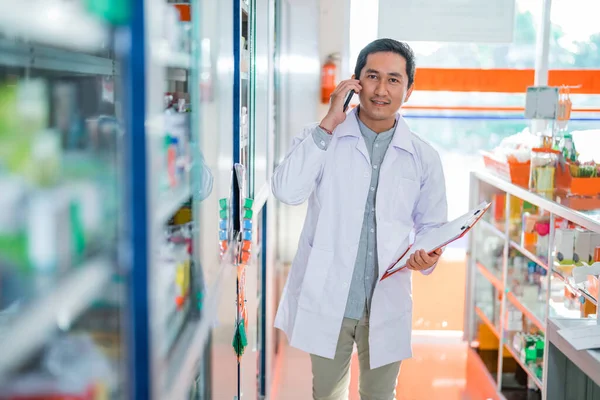 male pharmacist in uniform taking call and holding clipboard while looking at camera in pharmacy