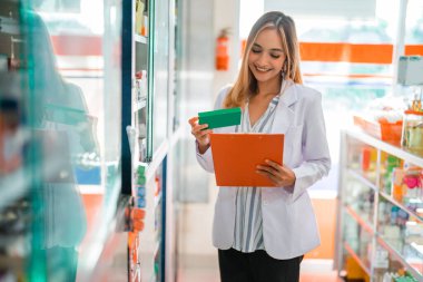Kadın eczacı elinde ilaç paketi, içindekileri kontrol ediyor ve eczanede pano tutuyor.