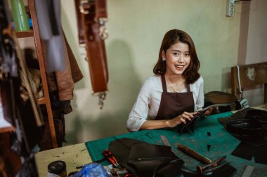 Kadın zanaatkar kameraya gülümserken bronzlaşmış deri parçaları tutuyor, deri önlük yapıyor.