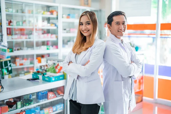 Smiling female pharmacist and male pharmacist with crossed hands standing inside drugstore