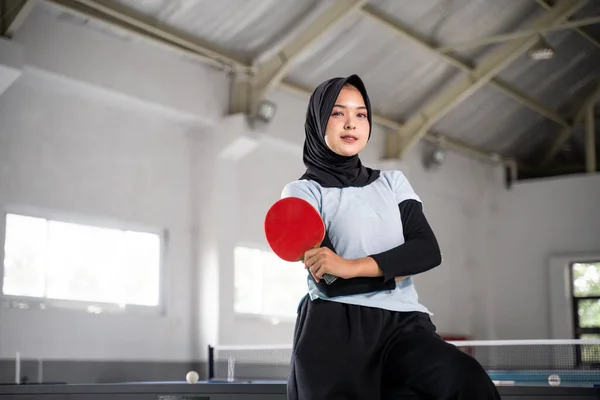 table tennis athlete in hijab holding paddle with crossed hands inside building