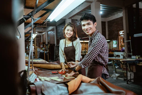 Asian male and female leather worker smiling while standing in his studio