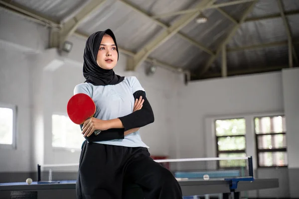 female table tennis athlete in hijab holding bat with crossed hands inside building