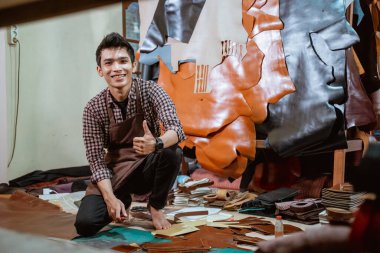 Atölyede çömelirken kameraya gülümseyen bir zanaatkar.