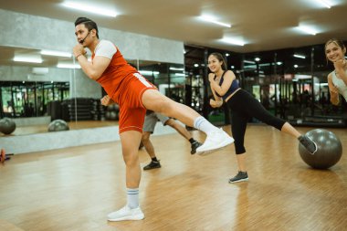 Erkek eğitmen, spor salonundaki beden eğitimi eğitimcileriyle yan yana bacak kaldırıyor.