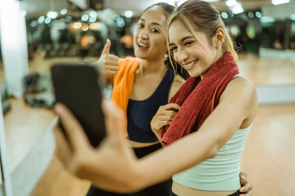 two happy asian women taking selfies together using a cellphone camera after a workout at the gym