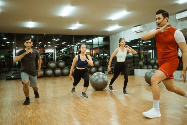 Spor salonunda jimnastik yapan bir grup genç. Arka planda jimnastik topu var.