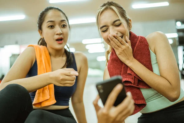 two asian women excited while using mobile phones together after a workout at the gym