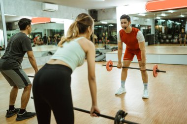 Erkek eğitmen, spor salonundaki bir grup gençle sırt kaslarını çalıştırmak için halterleri kaldırdığını gösteriyor.