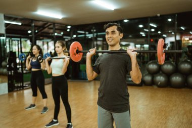 Genç Asyalı adam spor salonunda spor yaparken halteri göğsünün önüne kaldırıyor.
