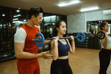 Erkek eğitmen, genç bayana halter kullanarak uygun hareketleri öğretiyor.