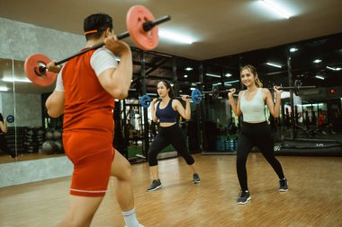 Eğitmen ve iki çekici kadın spor salonunda halterli çömelme hareketleri yapıyorlar.