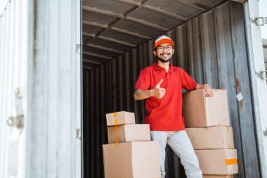 delivery man in red uniform with thumbs up standing between several boxes in container truck