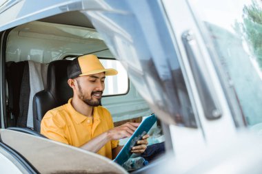 delivery man in yellow uniform using clipboard before delivering goods while sitting driving goods truck