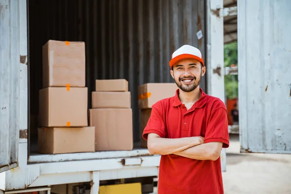 delivery man in red smiling with arms crossed standing against a background of a container full of boxes