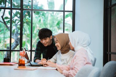 three young Asian people using a laptop working together in a cafe against a background of glass windows