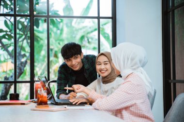 three young asian people discussing doing assignment together in cafe with glass windows in background
