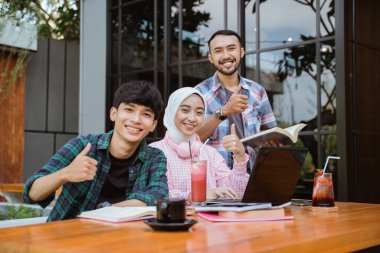 three smiling Asian students with thumbs up while studying together in cafe