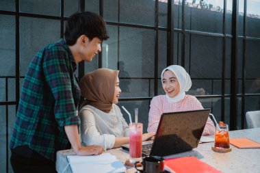 three young Asian people using a laptop and chatting in a cafe with glass windows in the background