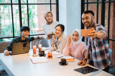 five young asian people smiling taking selfie using cell phone in cafe