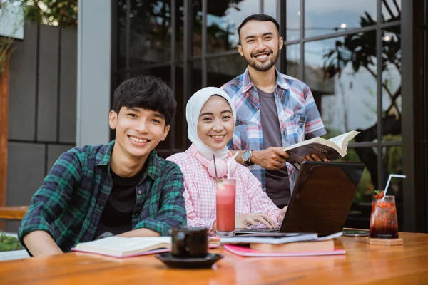 three smiling Asian students looking at camera while studying together in cafe