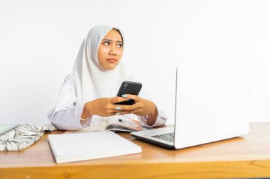 veiled high school student at desk with laptop thinking while using mobile phone on isolated background