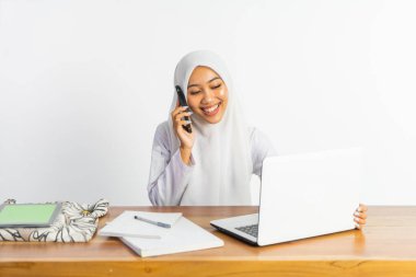 veiled high school student at desk with laptop while receiving cell phone call on isolated background