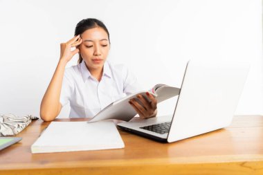 high school girl sitting at desk with laptop and holding book with thinking expression on isolated background