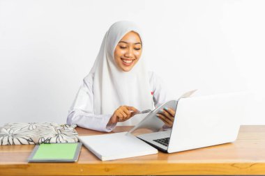 high school veiled girl sitting at desk reading book while using laptop on isolated background