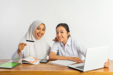 two smiling high school girls sitting at desk with laptop while showing blank cards on isolated background