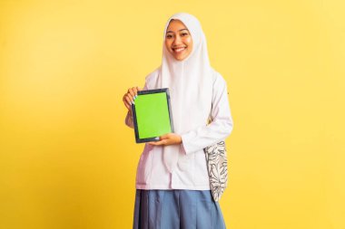 Smiling veiled high school student girl carrying bag showing blank screen a pad standing on isolated background