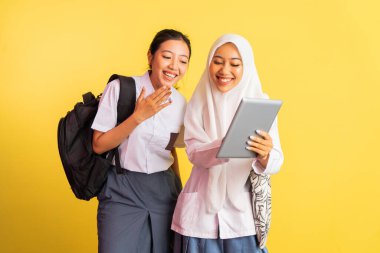 two high school girls carrying bags laughing while using digital tablet on isolated background